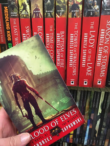 a hand holds a copy of Andrzej Sapkowski's book "Blood of Elves" in front of a row of his books